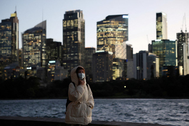 2025年7月10日、オーストラリアのシドニーで、中央ビジネス地区(CBD)のスカイラインを背景に、女性が夕暮れ時のウォーターフロントを歩いている。REUTERS/Hollie Adams