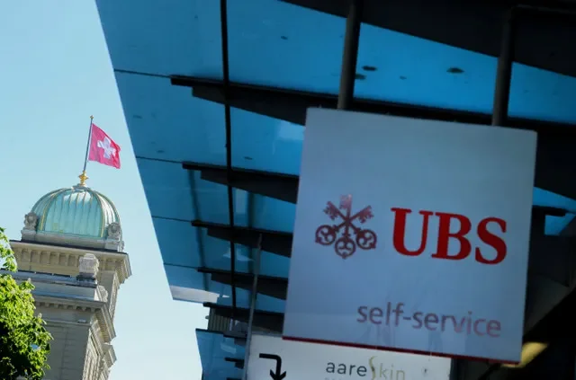 โลโก้ของ UBS หน้าธงชาติสวิตเซอร์แลนด์บนอาคารรัฐสภาสวิส (Bundeshaus) ก่อนการแถลงข่าวเกี่ยวกับข้อกำหนดเงินกองทุนของธนาคารสวิสในกรุงเบิร์น ประเทศสวิตเซอร์แลนด์ เมื่อวันที่ 22 เมษายน 2026 ภาพโดย REUTERS/Denis Balibouse/File Photo